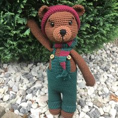 a crocheted teddy bear wearing overalls and a hat is standing in front of a bush