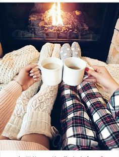 two people are holding mugs while sitting in front of a fireplace with their feet up