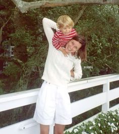 a woman holding a small child on her shoulders while standing next to a white fence