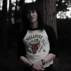 a young man standing next to a tree in the woods wearing a hellfire club t - shirt