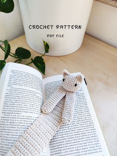 a crocheted cat laying on top of an open book next to a potted plant