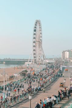 a large crowd of people walking and riding bikes on the beach next to a ferris wheel