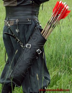 a man in black outfit holding red arrows with his back turned to the camera while standing in tall grass