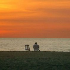 a man sitting on top of a bench next to the ocean under a colorful sky