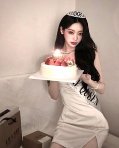 a woman in a white dress holding a cake with lit candles on it while wearing a tiara