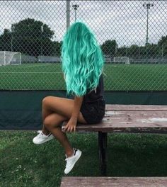 a woman with blue hair sitting on a bench in front of a fence and soccer field