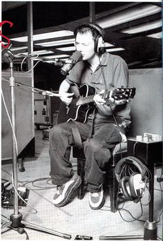 a man with headphones on sitting in front of microphones and playing an acoustic guitar