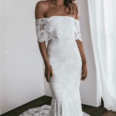 a woman standing in front of a window wearing a white dress with off the shoulder sleeves