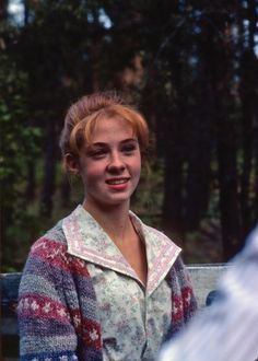 a woman sitting on top of a wooden bench next to a forest filled with trees