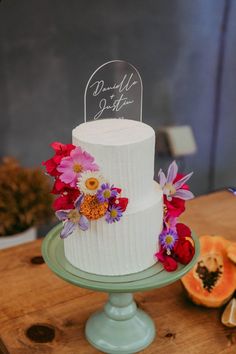 a three tiered cake with flowers on the top is sitting on a table next to sliced fruit