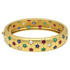 A vintage multi colour gemstone and yellow gold bangle bracelet by Cartier Paris, circa 1990. The 18ct yellow gold bangle has a rounded profile with a combination of diamonds, rubies, sapphires and emeralds all the way around, each in a five point star setting. The bangle is hinged to the side and secured with a concealed tongue and box clasp and safety catch. Maker Cartier Period circa 1980/90s Origin Paris Gemstones 12 x round brilliant cut diamonds - estimated 1.20ct 12 x sapphires - estimate Cartier Gold Bracelet With 17 Jewels, Cartier Yellow Gold Bangle, Cartier Yellow Gold Round Bangle, Cartier Gold Bangle With 17 Jewels, Vintage Cartier Watch, Five Point Star, Star Setting, Cartier Vintage, Yellow Gold Bangle