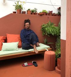 a woman sitting on top of a wooden bench next to potted plants and pillows