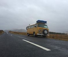 an old yellow van with a surfboard on the roof driving down a rural road