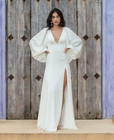 a woman standing in front of a wooden wall wearing a white dress with long sleeves