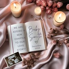 an open book sitting on top of a pink blanket next to two candles and flowers