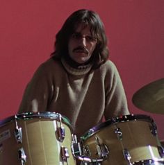 a man with long hair playing drums in front of a red wall and wearing a brown sweater