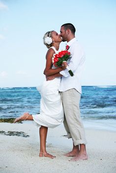 a bride and groom kissing on the beach in front of the ocean with red flowers