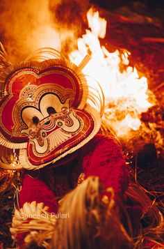 a person wearing a mask sitting in front of a fire with flames coming out of it