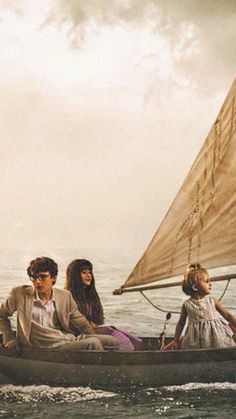 three people in a small boat on the water with a sailboat behind them,