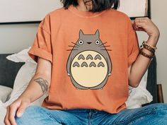 a person sitting on a bed wearing an orange tshirt with a totoro drawn on it