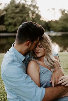 a man and woman cuddling in the grass next to a lake at sunset with their arms around each other