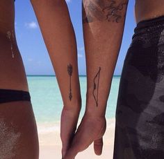 two people holding hands with tattoos on their arms near the ocean and sand in front of them