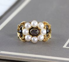 An immaculate antique Georgian mourning ring, crafted in 18k yellow gold with a lustrous split pearl halo around the center compartment which holds a momento of woven hair under crystal. The shoulders are delicately articulated in a baroque scroll design, accented by glossy black Victorian Yellow Gold Pearl Ring With Rose Cut Diamonds, Antique Cabochon Pearl Ring For Anniversary, Antique Pearl Ring With Cabochon For Formal Events, Antique Pearl Ring With Cabochon For Formal Occasions, Formal Heirloom Pearl Ring With Cabochon, Formal Heirloom Cabochon Pearl Ring, Antique Pearl Ring With Cabochon For Anniversary, Victorian Oval Cabochon Pearl Ring, Victorian Cabochon Pearl Ring Gift