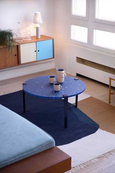 a blue table with two mugs on it in front of a couch and window