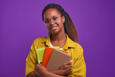 a woman in glasses is holding books and smiling at the camera while standing against a purple background