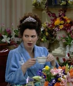 a woman sitting at a table with a tea cup in her hand and wearing a tiara