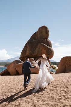 a bride and groom walking on the beach with large rocks in the background at their wedding