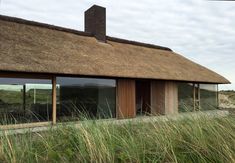 a thatched roof house with large windows and grass growing in the foreground on a cloudy day