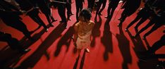 a woman walking down a red carpet in front of a group of people with their shadows on the floor