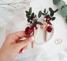 a person holding flowers on top of a white plate next to two wedding rings and greenery