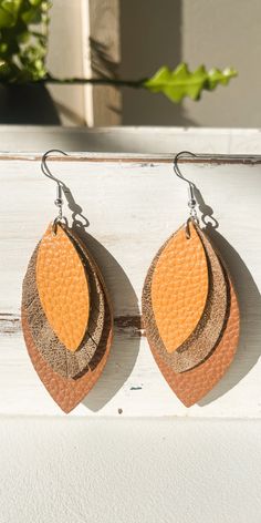 two pairs of leather earrings hanging from hooks on a white surface with plants in the background