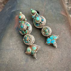 This old pair of akor earrings is made of tribal silver inlaid with turquoise in varying tones, from the bright blue inlaid into one of the lotus bud symbols at the bottom to the green at the top of that same earring. These earrings are old enough that the turquoise in some cases has been discolored, the spaces between stones dark and almost grimy looking. These are likely at least 70 years old and can be much older, as this style of earring would have been worn by women in the Lhasa region of Tibet, up until about the mid-20th century. They were generally hung at either side of the face hiding both ears and facing forwards to show their inset stones. These earrings are heavy, although not as heavy as antique sterling silver versions, which might weigh up to two or three times more per ear Traditional Turquoise Jewelry For Ceremonial Occasions, Turquoise Jewelry With Matching Earrings For Festivals, Traditional Blue Pendant Earrings, Traditional Silver Earrings With Inlay, Silver Spiritual Earrings For Rituals, Traditional Turquoise Drop Earrings, Traditional Silver Multi-stone Earrings, Traditional Turquoise Sterling Silver Earrings, Traditional Green Multi-stone Earrings