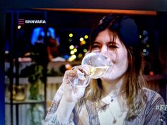 a woman drinking from a wine glass in front of a tv screen with the news on it