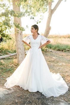 a woman in a wedding dress standing next to a tree with her hands on her hips