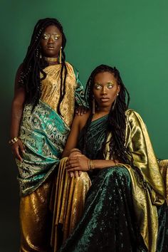 two black women in gold and green dresses sitting on a chair together, one is holding her arm around the other's shoulder