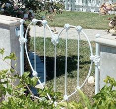 a white iron bed frame sitting in the grass next to a fence and bushes with pink flowers