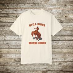 Check out my funny new wild west rodeo cowboy graphic T-Shirt, Done in the Retro Style of the 80s and 90's, this graphic western shirt, pokes gentle fun at the manly and bravery it takes to ride a bucking bronc horse. Every now and then you have to step back and say wow! Look at that guy ride! and wonder... How many rocking horse the went through as a toddler... ☺️??? Gotta love a cowboy! This super comfy comfort color tee has a relaxed fit and the cotton is soft and durable. This will make a great gift for the guy with a sense of humor! Comfort Colors Tshirts: About the Designer of FlooredByArt Studios: ☀️As an artist I try to capture the beauty of nature and the spirit of beloved animals with these stunning archival quality prints. Each design is crafted using cutting-edge digital artist Western Style Relaxed Fit Graphic T-shirt, Western Style Relaxed Fit T-shirt With Graphic Print, Western Style Crew Neck T-shirt With Graphic Print, Western Style Relaxed Fit T-shirt With Screen Print, White Graphic Print T-shirt For Western-themed Events, Casual White T-shirt For Western-themed Events, Western Style Screen Print T-shirt For Western-themed Events, Cotton Graphic Print Shirt For Rodeo, Casual Crew Neck T-shirt For Western-themed Events