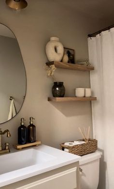 a white toilet sitting under a bathroom mirror next to a sink and shelf with items on it