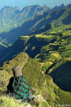 a man sitting on top of a lush green hillside next to a valley filled with mountains