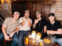 three men sitting at a table with drinks and pizzas in front of them, giving the peace sign