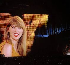 a woman with long blonde hair is smiling in front of a large screen that has an image of a man on it