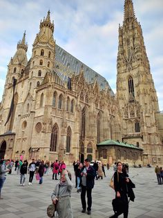 many people are walking around in front of a large building that has two towers on each side