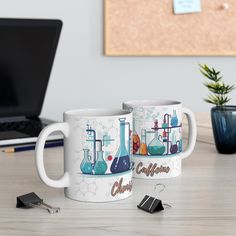 two coffee mugs sitting next to each other on a table with a laptop in the background