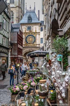 people are walking through an old town with lots of flowers on the sidewalk and buildings in the background