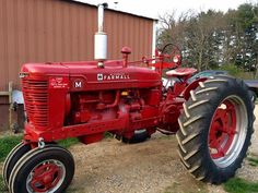 an old red farmall tractor is parked in front of a brown building and grass