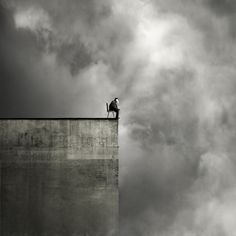 a man sitting on top of a cement wall next to a sky filled with clouds